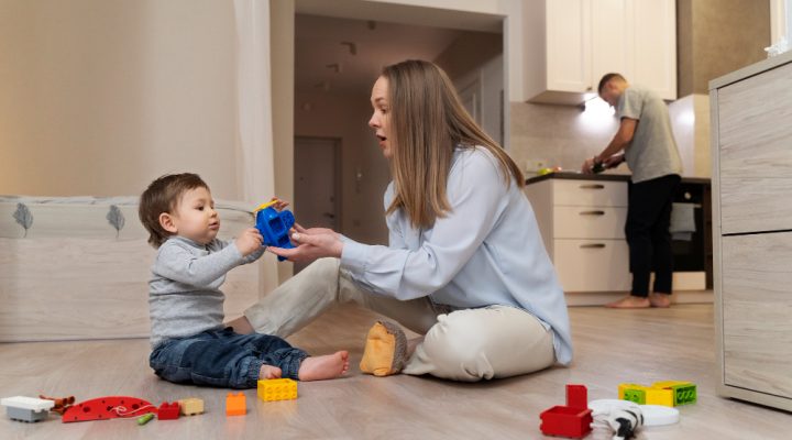 Mother playing with toddler while father cooks in background – assessing nanny training and family confidence.